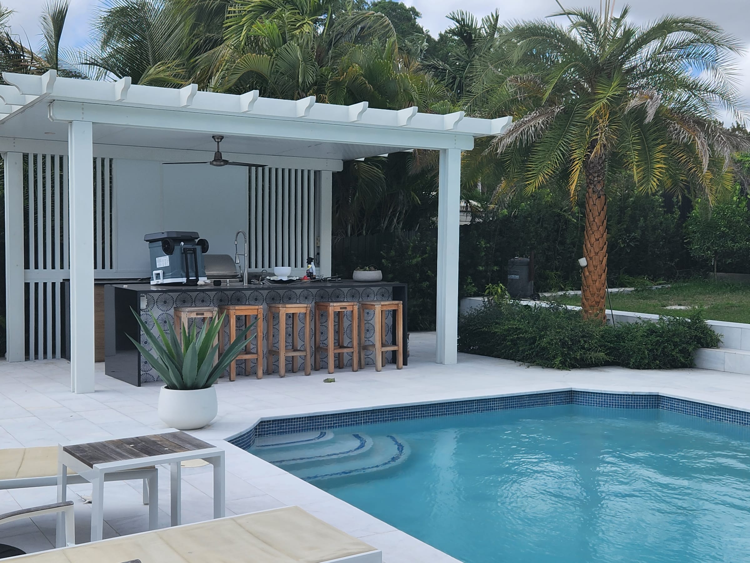Cocina de exterior con barra y banquetas bajo pérgola de aluminio blanco junto a la piscina