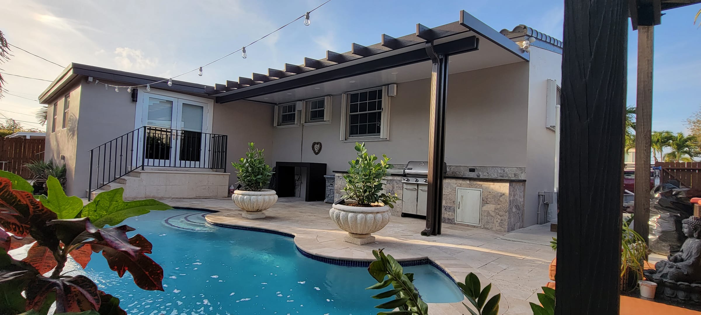 Cocina de exterior en piedra con parrilla bajo pérgola de aluminio bronce junto a la piscina